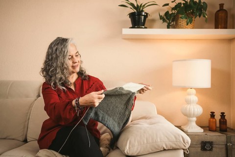 A woman with gray hair knits peacefully on a beige sofa, surrounded by plants and warm lighting.