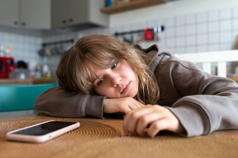 Teen girl rests her head on a table at home, deeply absorbed by her smartphone, highlighting her