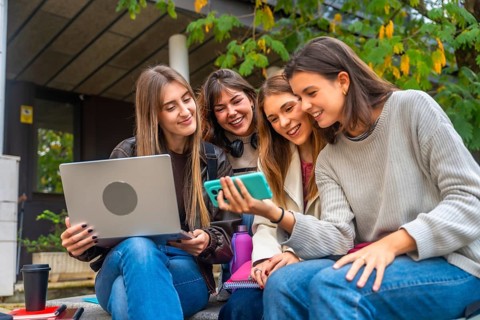 Group of female university students sitting outdoors, interacting with a smartphone and laptop,