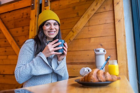 Woman holding a coffee mug, smiling at the camera, and enjoying a croissant, orange juice, and hot