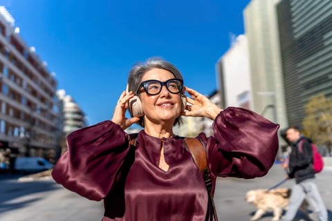 Mature woman with gray hair wearing headphones and eyeglasses, finding joy and relaxation while