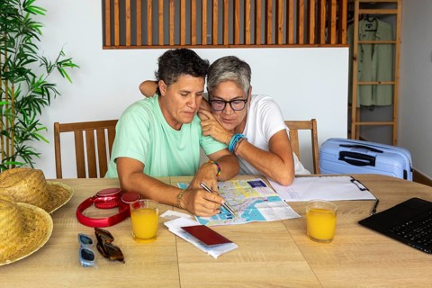 A lesbian couple, plans their summer holiday with enthusiasm. They sit at a wooden table,