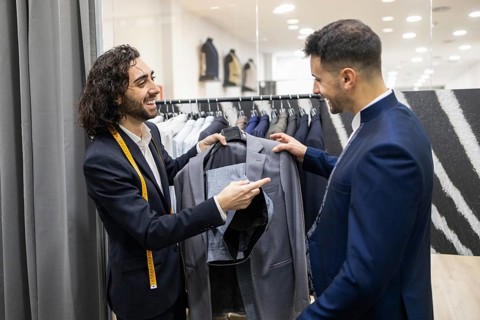 A tailor with a measuring tape assists a client in selecting a stylish suit from a rack in a suit