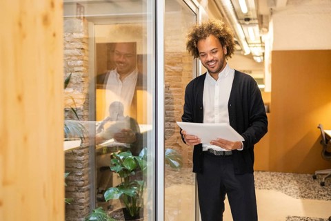 A man in a coworking space holds a document, smiling as he stands near a glass wall. The modern