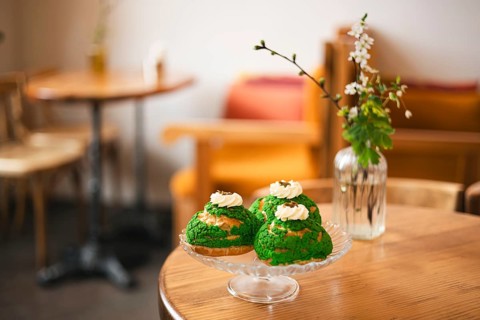 A glass plate with matcha choux pastries with whipped cream sits on a wooden table in a cozy cafe.