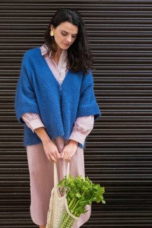 A woman wearing a blue cardigan and striped dress holds a mesh bag filled with fresh greens. Her