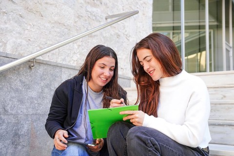 Two friends sitting on steps outdoors, one holding a bright green notebook and a pen, the other a