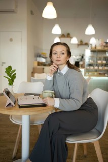 Woman sits at a cafe table with a coffee cup, tablet, and notebook. She appears thoughtful and