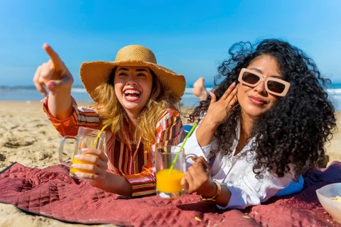Two cheerful female friends relaxing on a picnic blanket by the ocean, holding refreshing drinks