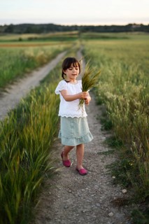 A young girl in a white blouse and green skirt walks a dirt path, holding stalks of wheat. The calm