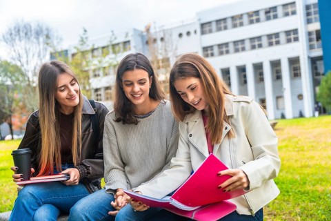 Three university students studying together outdoors on campus, sharing textbooks, coffee and notes