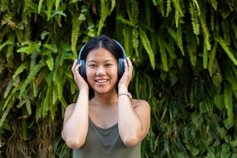 A young Chinese woman wearing headphones enjoys music against a lush green urban background. Her