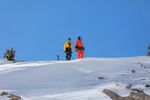 Two snowboarder friends in colorful winter gear stand on a snowy mountain slope, ready to descend.