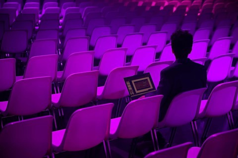 A indian man with a laptop sits amidst rows of empty chairs illuminated by purple lighting at a