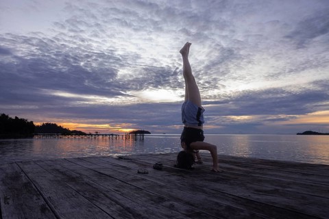 A tourist performs a headstand yoga pose on a wooden pier during a stunning sunset on Waigeo