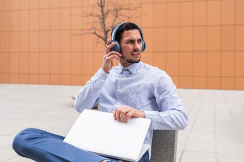 Man in business casual attire sits outside with a laptop and headphones, appearing focused while