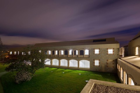 A modern hospital with stone facade and arched windows is lit from within against a twilight sky.
