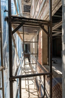 Sunlit scaffolding lines the narrow walkway of a construction site, flanked by exposed brick walls.