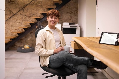 A young man sits casually in a modern coworking space, holding a coffee cup. A tablet displays