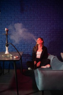 A woman is sitting on a sofa, calmly enjoying a hookah with smoke swirling in a cozy lounge. She is