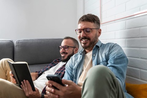 A joyous gay couple relaxes on the couch, one reading a book and the other using a smartphone,
