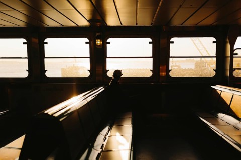 A lone traveler sits on a ferry, silhouetted against a warm sunset in New York. The light filters