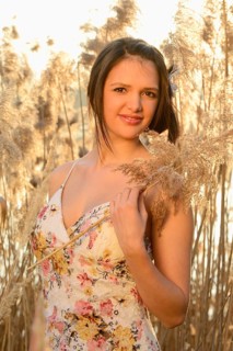 A woman in a floral dress holding a plant and smiling amongst reeds in the serene evening light,