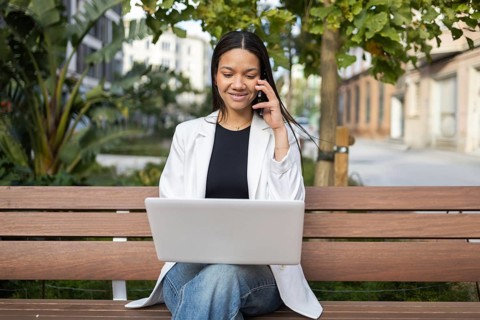A professional woman in casual attire sits on a bench, working on her laptop while talking on the