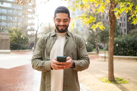 Smiling man using a smartphone in a park of the city, surrounded by autumn leaves and gentle rain.