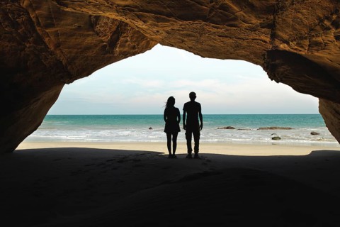 A couple stands inside a cave at Mancora Beach, Peru, gazing at the ocean. The cave's entrance