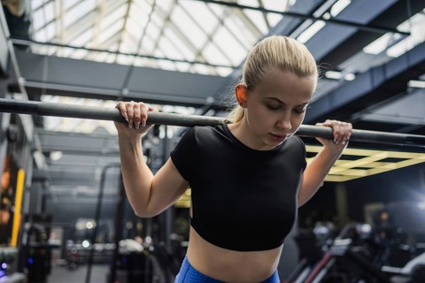 A focused blonde woman lifting a barbell in a contemporary gym. She wears a black crop top and blue