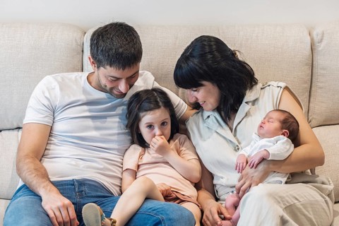 A young family sits together on a couch, radiating joy. The mother cradles a newborn baby, while