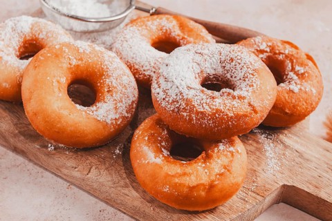 A wooden board holds several freshly made donuts. The donuts are round and have powdered sugar on