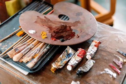 A wooden palette with mixed colors rests on a case of paintbrushes, with used paint tubes nearby.