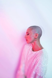 A middle-aged woman with a shaved head poses elegantly, wearing a sparkling top. The soft pink