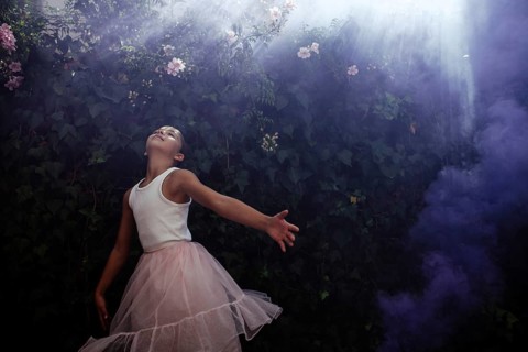 A teenage girl in a pink tulle skirt dances gracefully under the foliage, basking in the sunshine