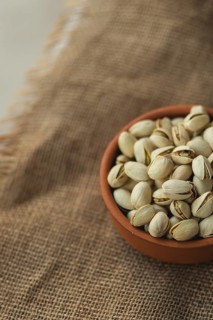 A terracotta bowl filled with pistachios rests on a rustic burlap fabric. The image highlights the