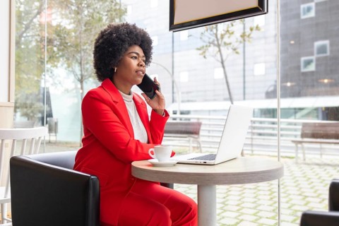A professional woman in a red suit sits in a modern cafe, talking on her phone while working on a