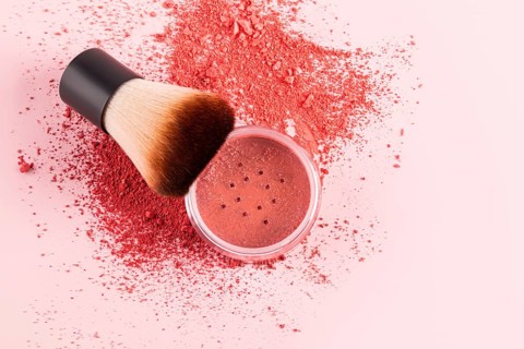 A compact blush sits on a pastel pink surface with a makeup brush. The soft bristles are ideal for