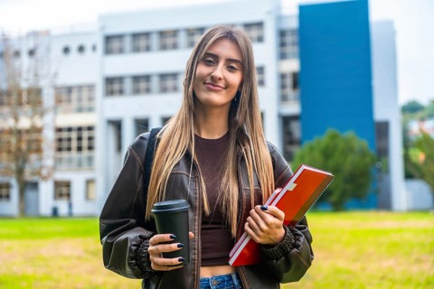 Young female student smiling and standing on a university campus, holding a takeaway coffee cup and