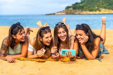 Young women friends lying on a towel on warm golden sand, laughing and watching content on a