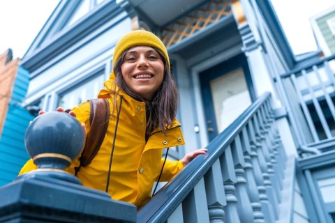 Smiling woman wearing a yellow raincoat and beanie, stepping up blue victorian house stairs during