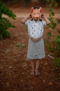 A young child stands in a vineyard, playfully holding a pig mask over their head. The scene