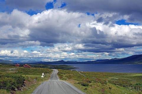A rural road passes by a lake surrounded by mountains and a dramatic sky, Fjell, Jotunheimvegen,