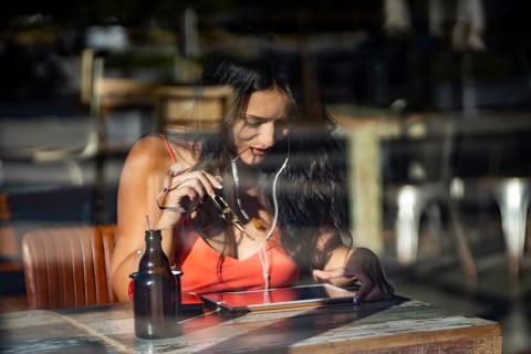 A woman sits in a cafe, absorbed in her tablet and wearing headphones. Sunlight streams in, casting