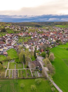 View of a village with gardens and fields, embedded in a green landscape under a cloudy sky,