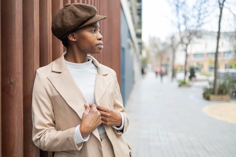 Stylish black woman in a coat and cap leans against a wooden wall on a city street Her thoughtful