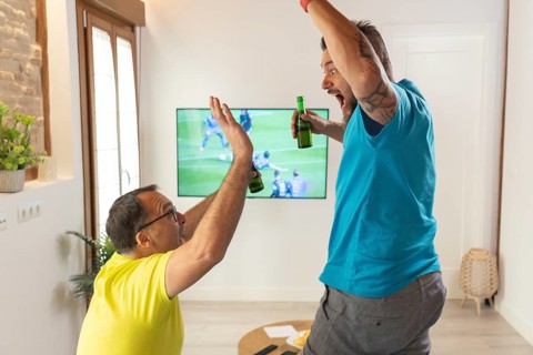 Two friends celebrating a goal with a high five while watching a soccer match on TV at home.