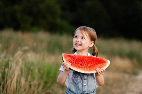 A cheerful child holding a slice of watermelon, enjoying the warm summer day outdoors in a field.