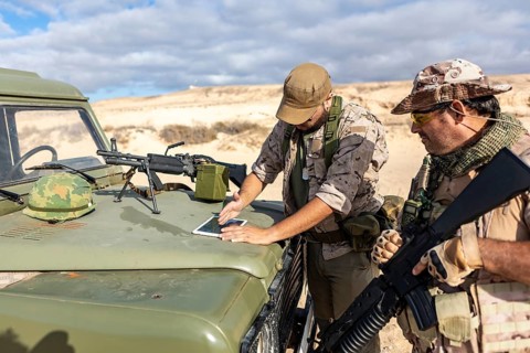 Two military soldiers in tactical gear analyze digital maps on a tablet atop a vehicle, preparing